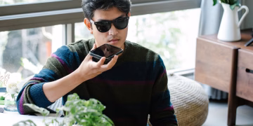 A man wearing sunglasses uses a smartphone's voice assistant in a cozy indoor setting.