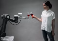 A woman in casual attire shares a toast with an advanced robotic arm, reflecting modern technology and innovation in robotics.