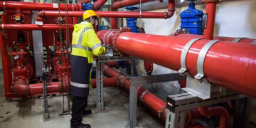 Engineer with safety gear inspecting red industrial piping system indoors.