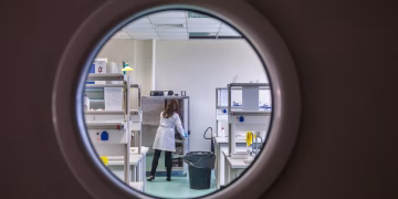 A female scientist in a lab coat working inside a state-of-the-art laboratory through a circular window view.