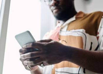 A close-up of a man using a smartphone by a window, featuring a modern patterned shirt.