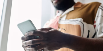 A close-up of a man using a smartphone by a window, featuring a modern patterned shirt.