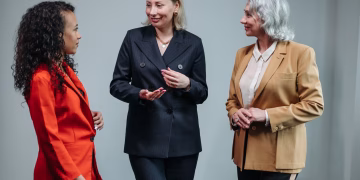 Three women in business attire having a conversation indoors, showcasing diversity and teamwork.