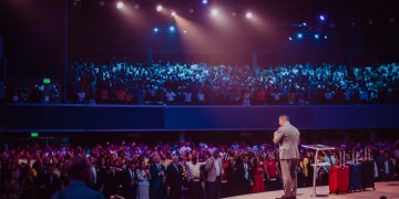 A speaker in a gray suit captivates a large crowd on stage during a motivational event.